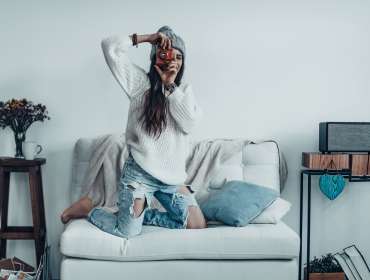 young woman in casual wear and knit hat taking a photo by her retro camera while standing on the sofa at home