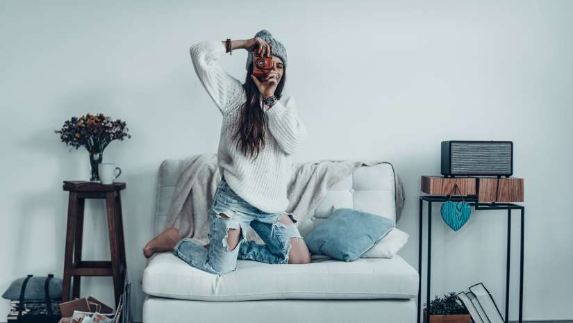 young woman in casual wear and knit hat taking a photo by her retro camera while standing on the sofa at home