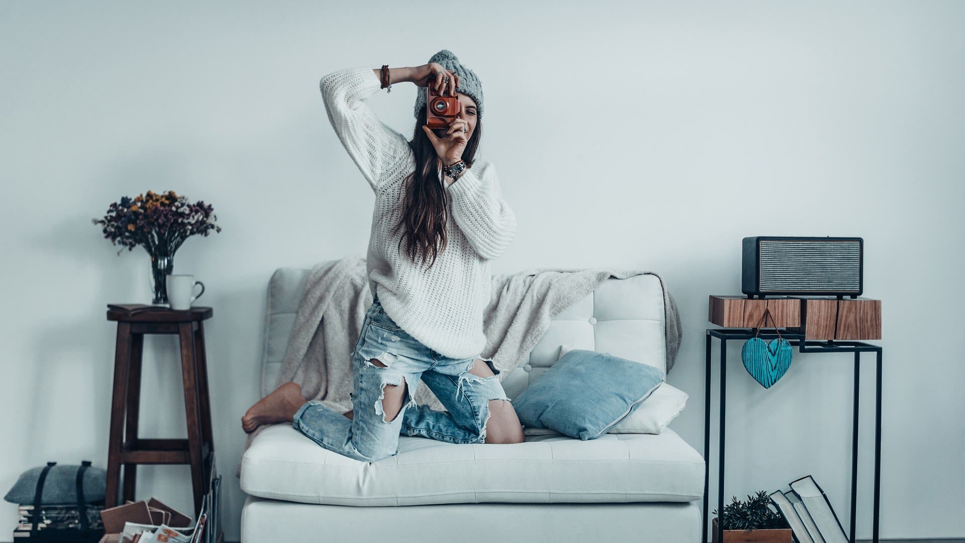 young woman in casual wear and knit hat taking a photo by her retro camera while standing on the sofa at home