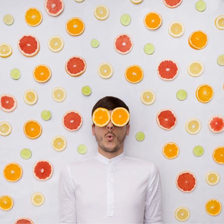 man covers eyes with sliced oranges with fruity background