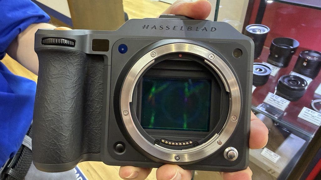 A person holding a large black Hasselblad medium format mirrorless camera body, showing the lens mount open to reveal a significantly large medium format camera sensor size with iridescent reflections.