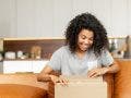 Positive young woman sitting on the couch with carton box on laps, feeling curious about ordered item from an online store, smiling client satisfied with fast courier delivery service