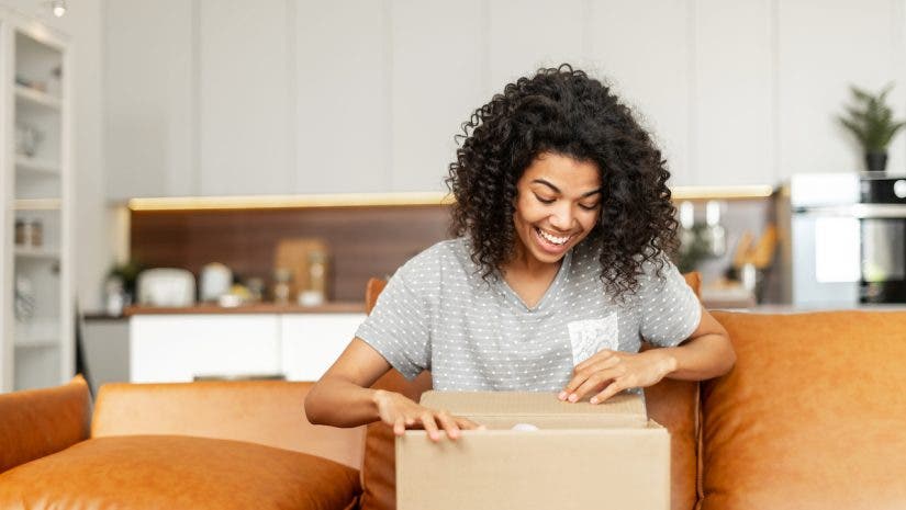 Positive young woman sitting on the couch with carton box on laps, feeling curious about ordered item from an online store, smiling client satisfied with fast courier delivery service