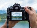 Hands of a male photographer holding a digital camera taking pictures of a idyllic landscape with a lake and mountains while the picture shows at the display