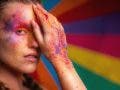 A colorful background reflects the colorful makeup on this female model's face and hands. She has one hand covering her eye.