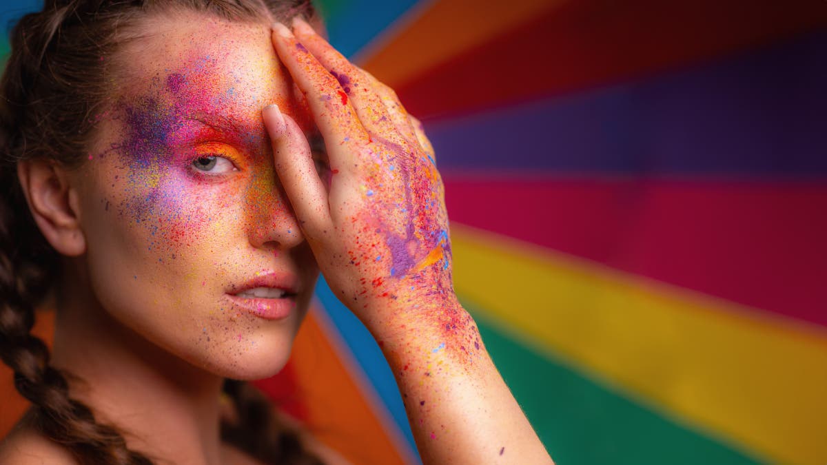 A colorful background reflects the colorful makeup on this female model's face and hands. She has one hand covering her eye.