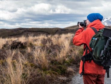 A man stands in a field making a photograph, wearing a camera backpack.
