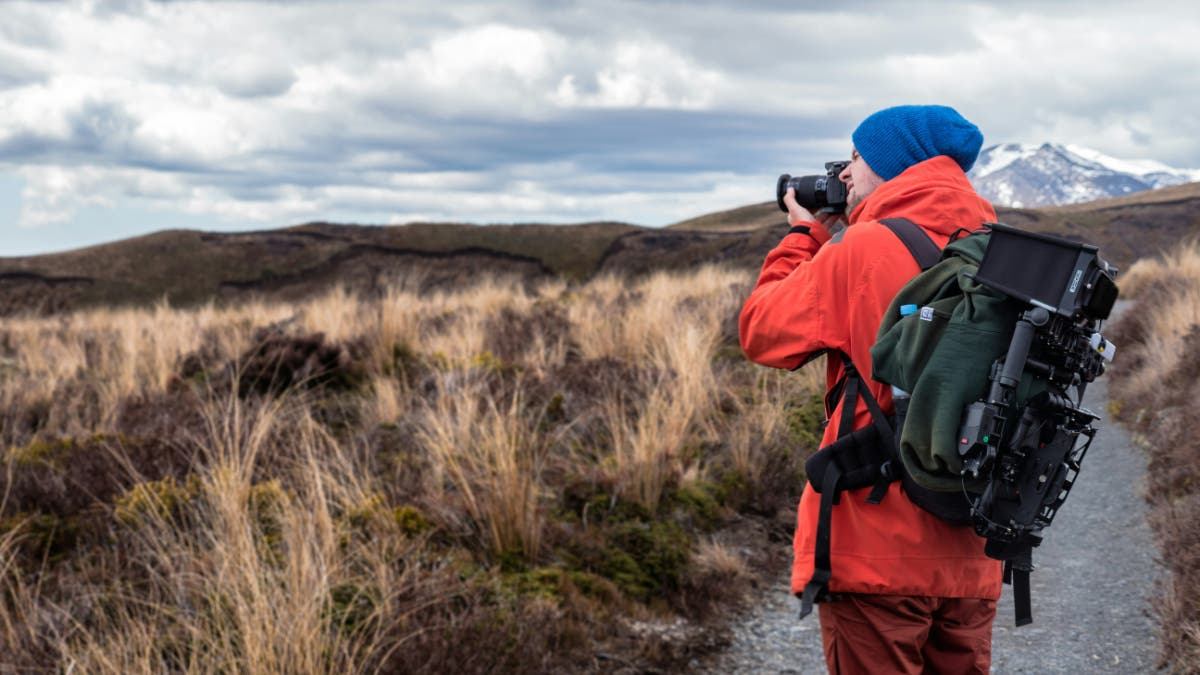 A man stands in a field making a photograph, wearing a camera backpack.
