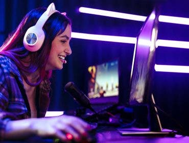 Happy young woman live streaming a video game, sitting in front of a multi monitor setup with a microphone for commentary and a headset for communication. Female engaging with fellow gamers on an online platform.