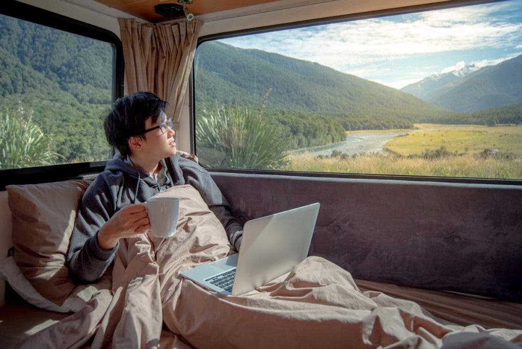 Young Asian man drinking hot coffee working with laptop computer on the bed in camper van with mountain scenic view through the window, digital nomad on road trip concept
