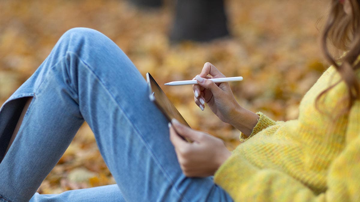 Young woman model sitting with tablet iPad Pro 2021 and painting with pencil stylus in autumn park with yellow foliage maple leaves.
