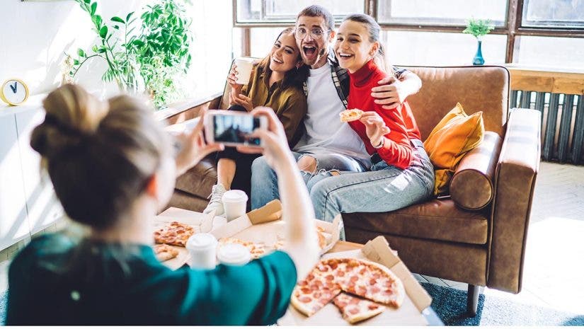 Cheerful smiling people embracing and sitting on couch by table with delivered pizza and cup while woman taking photo on smartphone in living room