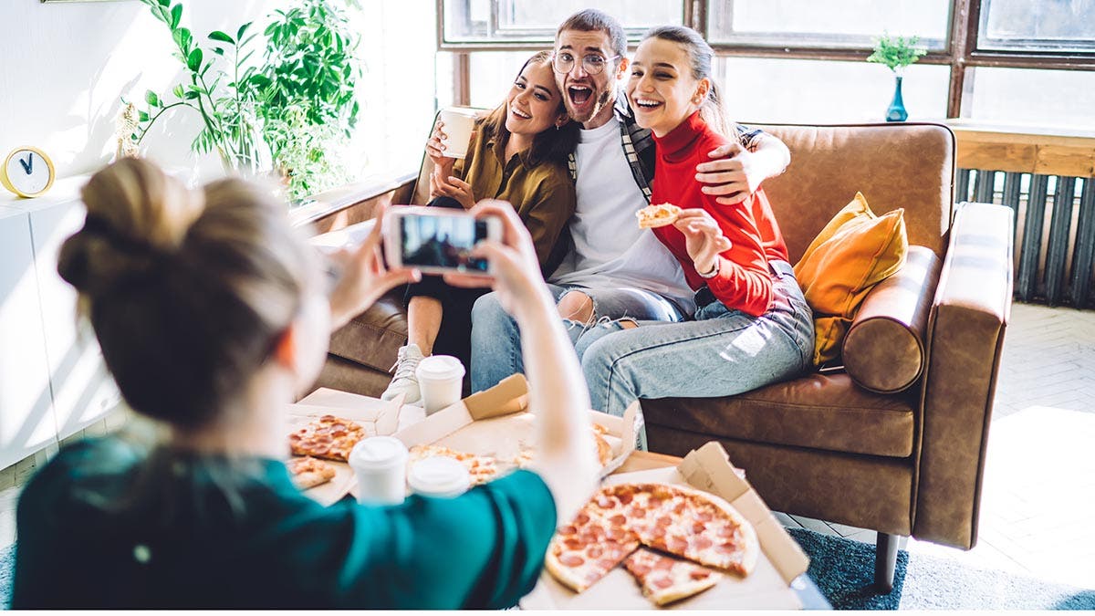 Cheerful smiling people embracing and sitting on couch by table with delivered pizza and cup while woman taking photo on smartphone in living room