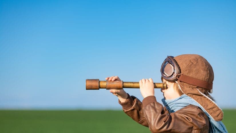 Happy child looking through spyglass outdoor in spring green field. Kid having fun against blue sky background. Imagination and freedom concept
