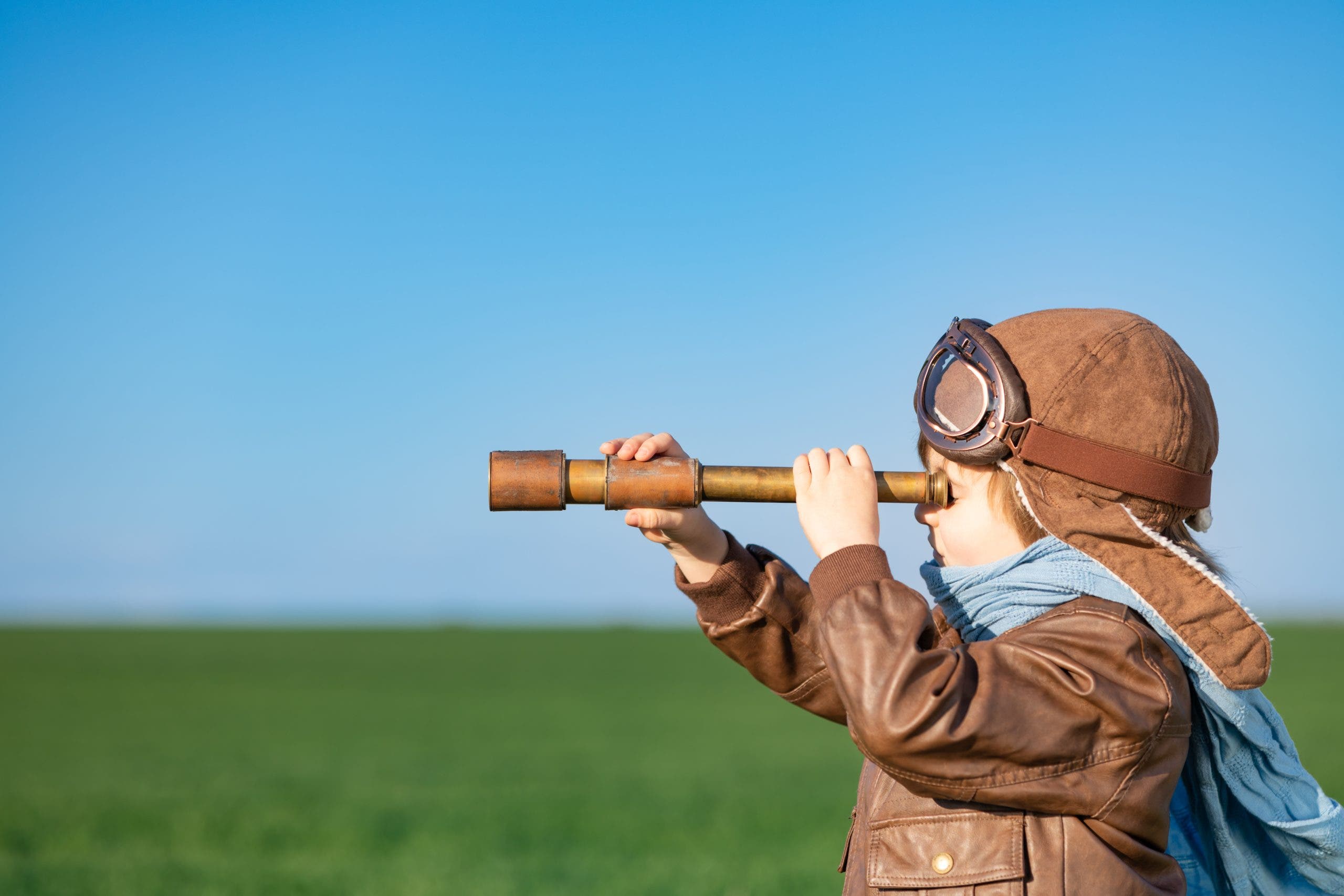 Happy child looking through spyglass outdoor in spring green field. Kid having fun against blue sky background. Imagination and freedom concept