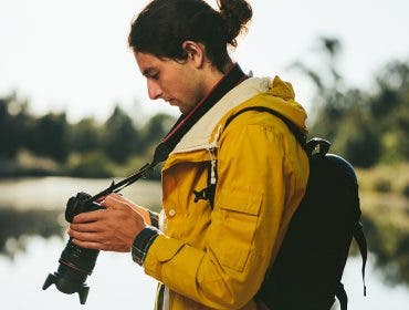 Side view of a tourist standing near a lake looking at his dslr camera. Close up of a traveler looking at his digital camera standing outdoors.