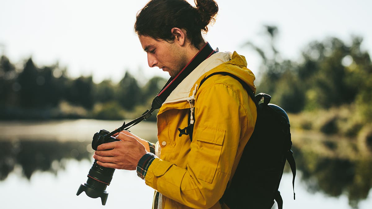 Side view of a tourist standing near a lake looking at his dslr camera. Close up of a traveler looking at his digital camera standing outdoors.