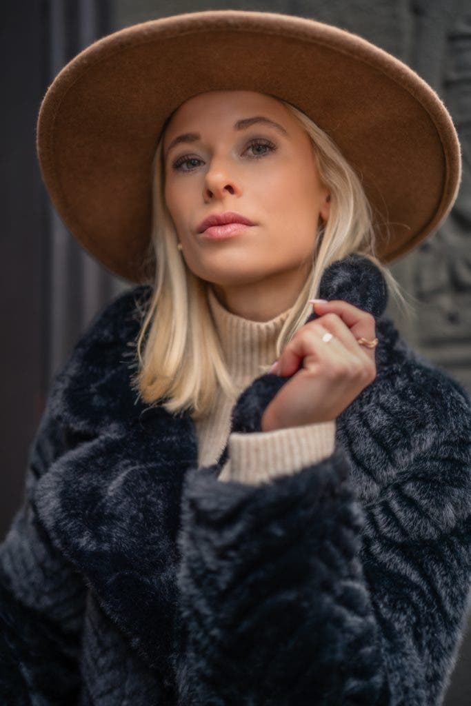 The photograph above prominently features the woman adorned in an elegant hat and a luxurious fur coat, making her the unmistakable focal point of the composition. The tight framing around her figure eliminates any distractions, while the shallow depth of field blurs the background, drawing the viewer's eye exclusively to her captivating presence.