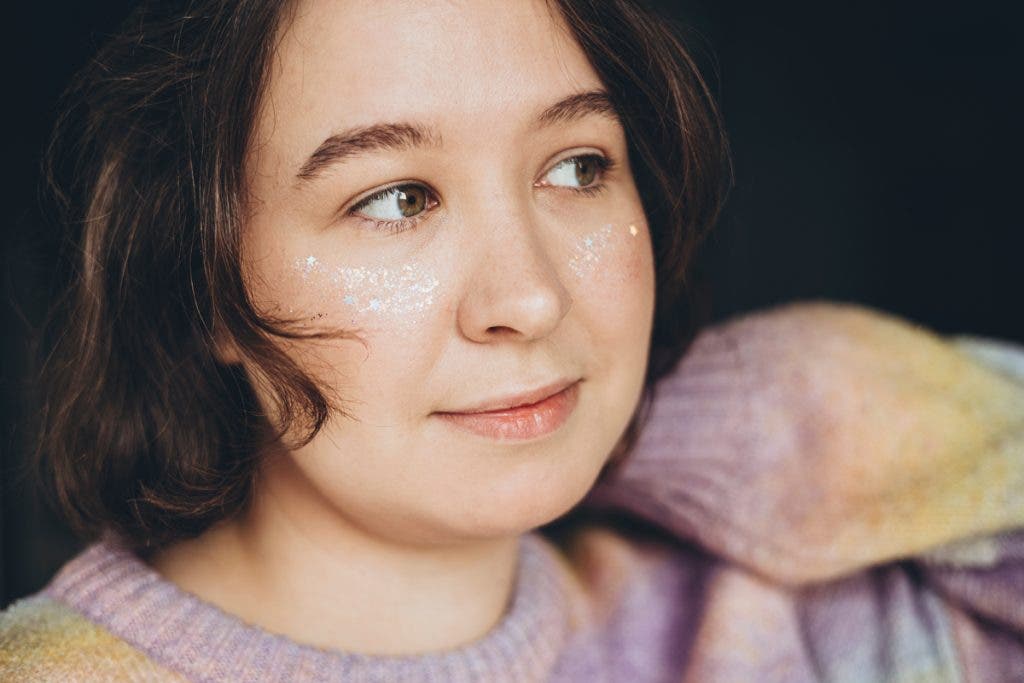 Close up natural colors portrait of beautiful girl with vivid and festive glitter makeup at home. Charming cheerful brunette with short wavy hair and sparkling makeup. Woman with shiny freckles.