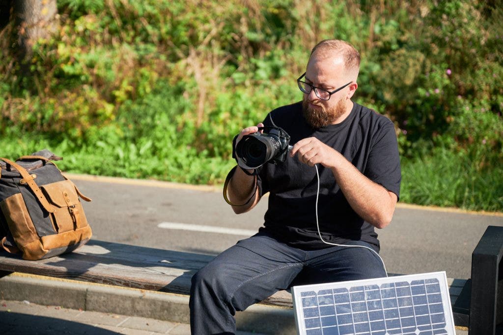 Bearded male photographer sits on bench, connecting camera to solar panel for charging. Integration of renewable energy into photography, using solar power to charge equipment during outdoor shoots.