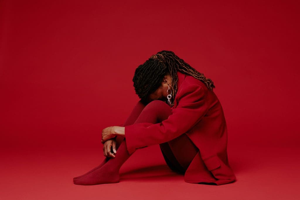 Woman with braided hair sitting on floor, wearing red jacket and pants against matching red background, facing downwards with contemplative pose