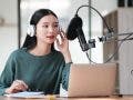 A woman wearing headphones is sitting at a desk with a laptop and a microphone. She is recording a podcast or a voiceover