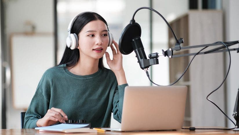 A woman wearing headphones is sitting at a desk with a laptop and a microphone. She is recording a podcast or a voiceover