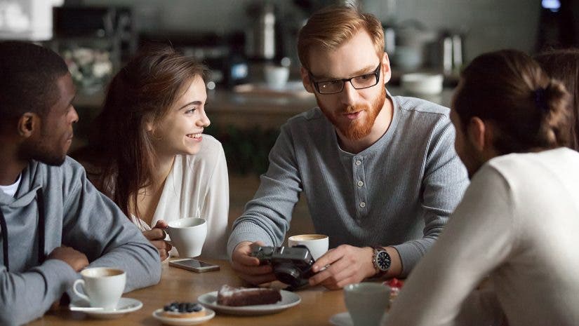Young redhead guy holding camera showing photos sharing recent memories or travel impression hanging with diverse friends, smiling multiracial millennial people talking sitting together at cafe table