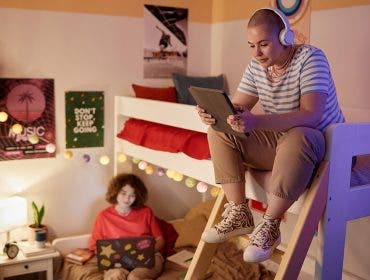 Portrait of students dorm room with bunk bed and two young women using tech devices, copy space