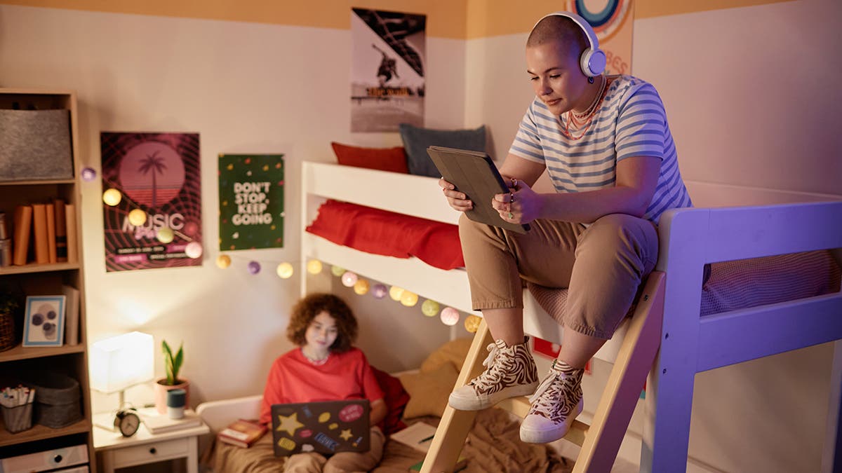 Portrait of students dorm room with bunk bed and two young women using tech devices, copy space