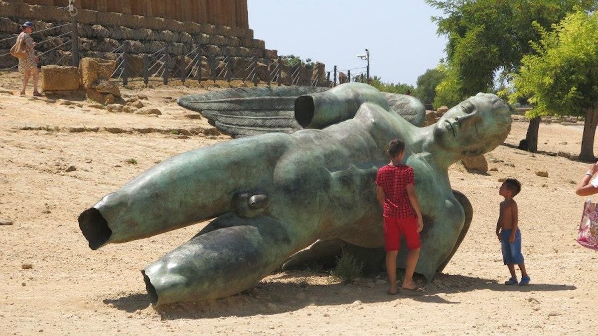 Children exploring a large bronze sculpture of a fallen figure at an archaeological site. The weathered green patina sculpture lies on sandy ground with ancient stone temple ruins visible in the background. A child in red shorts stands near the sculpture while another child observes from a distance. Trees and modern infrastructure are visible beyond the archaeological area.
