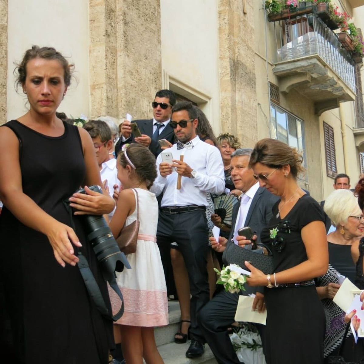 A crowd gathered on a narrow street outside a weathered building during what appears to be a wedding or formal ceremony. In the foreground, a woman in a black dress holds a professional camera with a large lens. Behind her, attendees in formal attire including suits and dresses mingle, with some holding drinks and phones. A young girl in a white dress with a pink sash stands among the adults. The building facade shows aged plaster walls with a balcony decorated with flowers visible in the upper right corner.