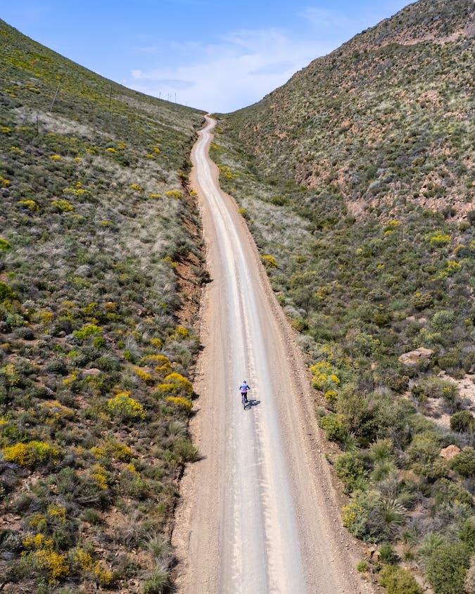 An aerial view showing a cyclist on a long dirt road, effectively using the road as a leading line and the cyclist to provide a powerful sense of scale against the expansive landscape.