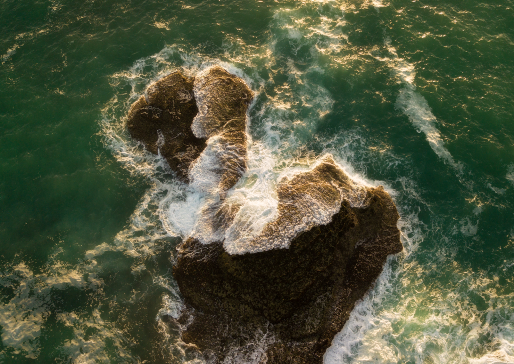 A unique aerial perspective captures a rock formation extending into the ocean, beautifully illuminated by soft sunrise light. The shot highlights the rock's texture, the glimmering water, and the white, creamy patterns of the waves.