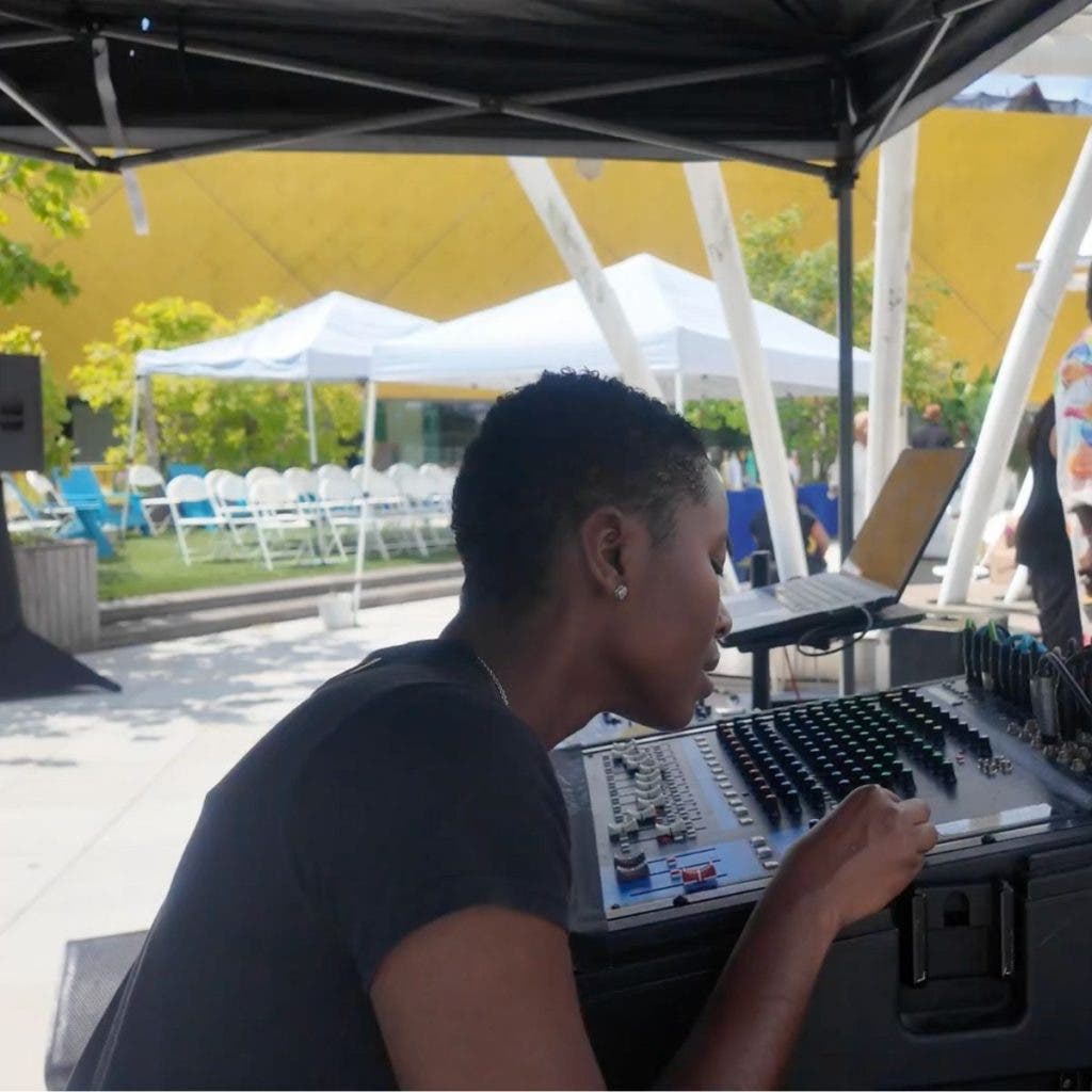 A production professional monitors a large analog audio mixing board under a tent, managing the sound for an outdoor production utilizing a two-camera setup.