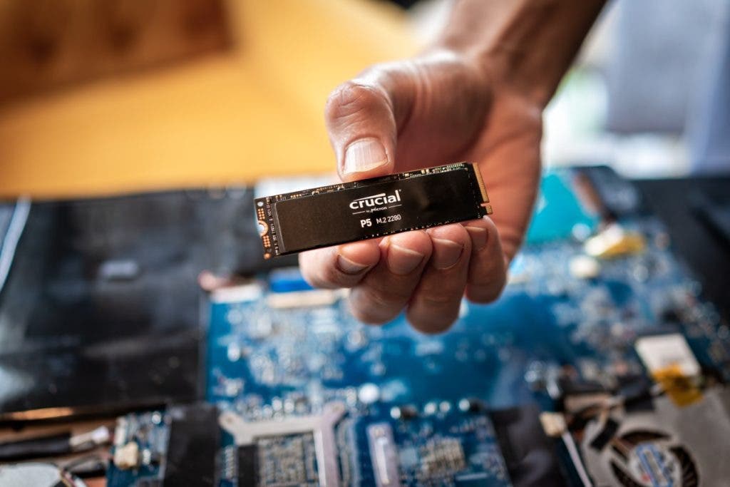 A person's hand holding a Crucial P5 M.2 NVMe SSD, illustrating a high-performance memory storage component held over an open laptop motherboard.