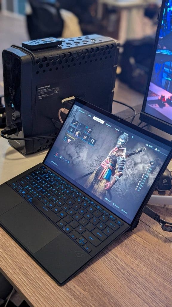 An angled shot of a gaming workstation highlighting a laptop running a character equipment screen from a video game, positioned next to a black CyberPower UPS