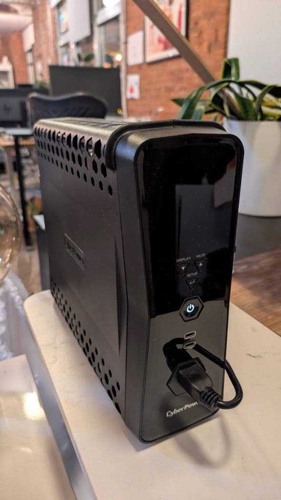 A sleek black CyberPower battery backup unit sits on a desk as part of a gaming workstation, featuring a glowing blue power button and multiple USB ports on the front panel.