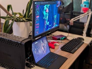 A comprehensive gaming workstation featuring a dual-monitor setup with a laptop serving as a secondary screen, an external mechanical keyboard, a red controller, and a large battery backup unit.