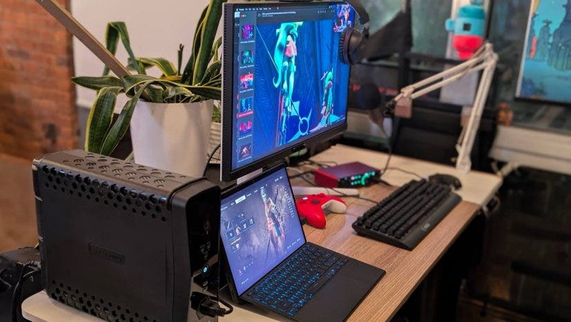 A comprehensive gaming workstation featuring a dual-monitor setup with a laptop serving as a secondary screen, an external mechanical keyboard, a red controller, and a large battery backup unit.