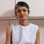 A portrait photograph of Shailee Jain Noronha, an Indian woman with short, curly, graying hair and yellow-rimmed glasses. She is wearing a light gray sleeveless top and small gold earrings, looking directly at the camera against a background of white modern cabinetry.