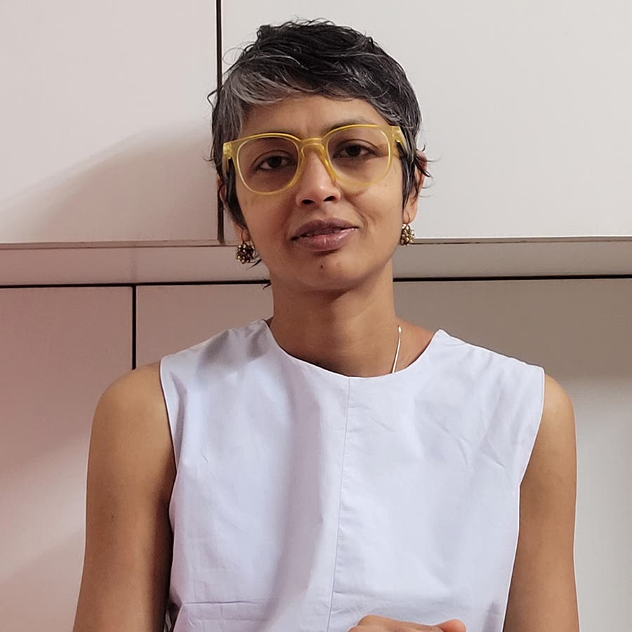 A portrait photograph of Shailee Jain Noronha, an Indian woman with short, curly, graying hair and yellow-rimmed glasses. She is wearing a light gray sleeveless top and small gold earrings, looking directly at the camera against a background of white modern cabinetry.
