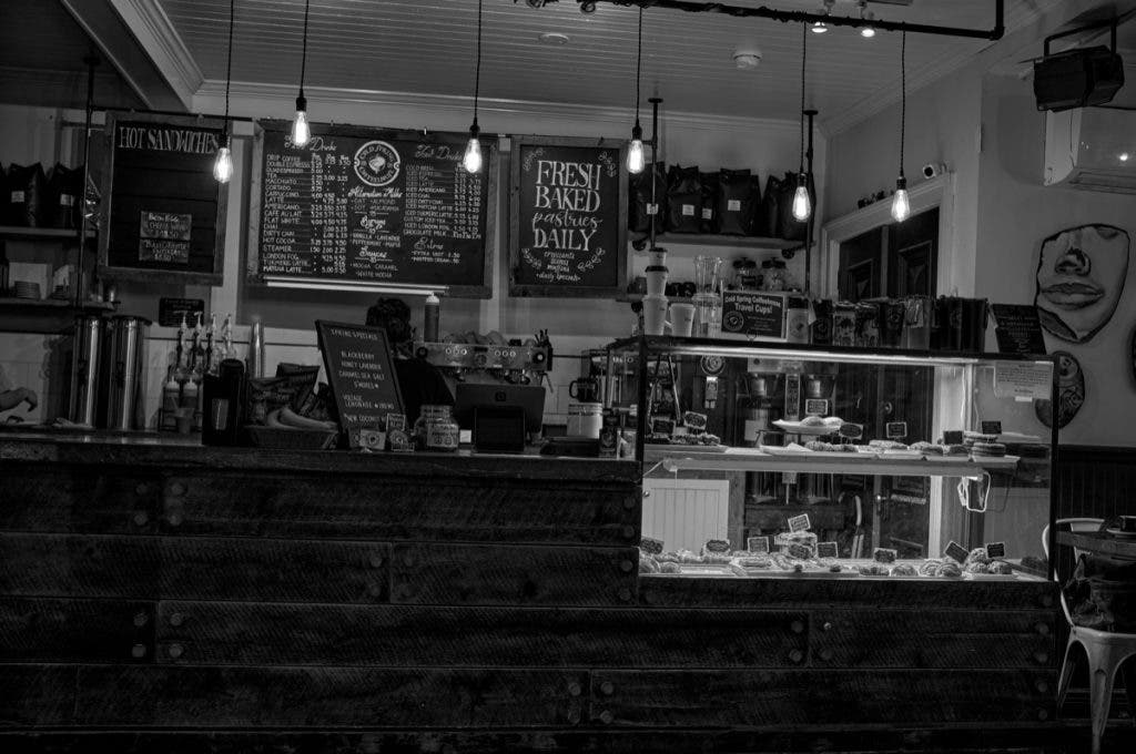 A high-contrast black and white interior of a rustic coffee shop featuring a dark wood counter, hanging Edison bulbs, and chalkboard menus. Shot by Daniel Norton on a Leica M11 Monochrom.