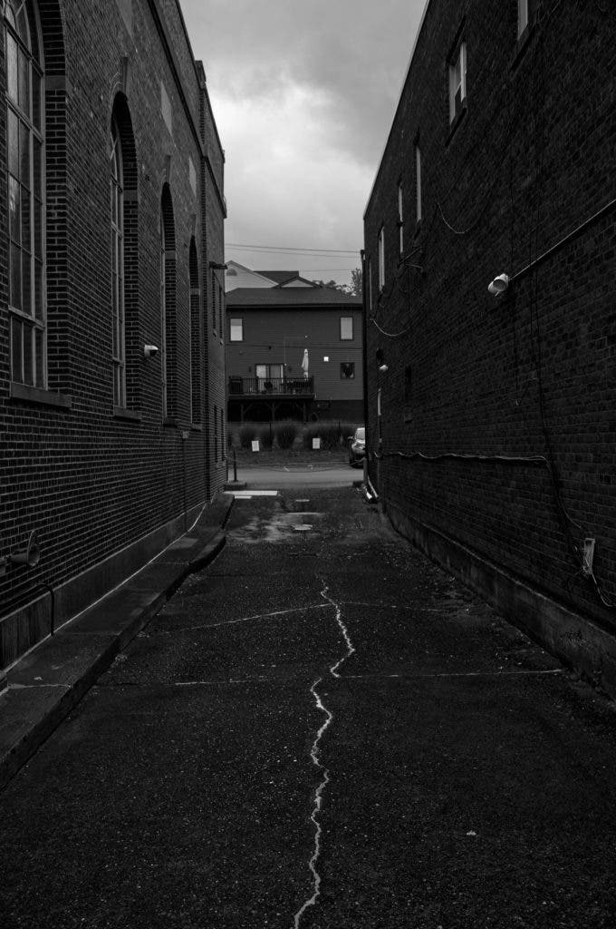 A moody, vertical black and white photograph looking down a narrow alley between two brick buildings toward a house in the distance, with a prominent crack running through the asphalt. Shot by Daniel Norton on a Leica M11 Monochrom.