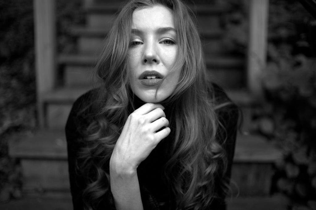 A close-up, shallow depth-of-field black and white portrait of a woman with long, wavy hair. The woman is Marisa Roper. She is looking directly into the camera with her chin resting on her hand, sitting on wooden outdoor steps. Shot by Daniel Norton on a Leica M11 Monochrom.