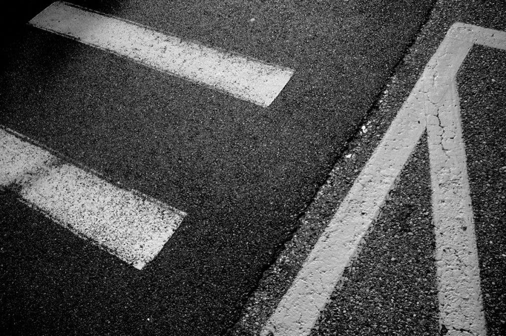 A black and white abstract top-down shot of textured asphalt. Thick, painted white road lines intersect and run parallel across the frame, highlighting the gritty texture of the pavement. Shot by Daniel Norton on a Leica M11 Monochrom.