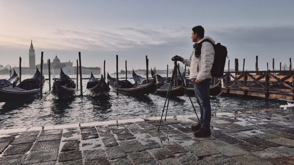 A man stands on a cobblestone pier in Venice, Italy, looking out over the water at dawn. He is positioned next to a camera mounted on a tripod and wears a large camera backpack. In the background, several gondolas are moored, and the silhouette of the San Giorgio Maggiore church is visible across the hazy lagoon.