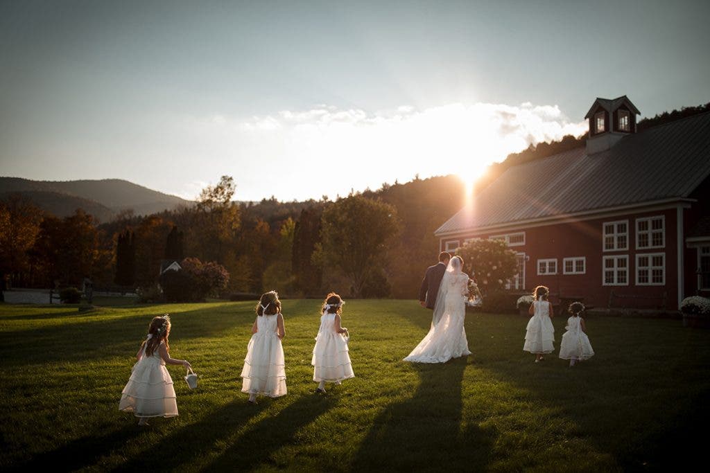 A wedding couple and four flower girls walking across a lush green lawn toward a red barn during a golden hour sunset, shot by Susan Stripling.