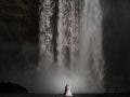 A wedding couple standing at the base of a massive, powerful waterfall, highlighting the epic scale of the landscape, shot by Susan Stripling.