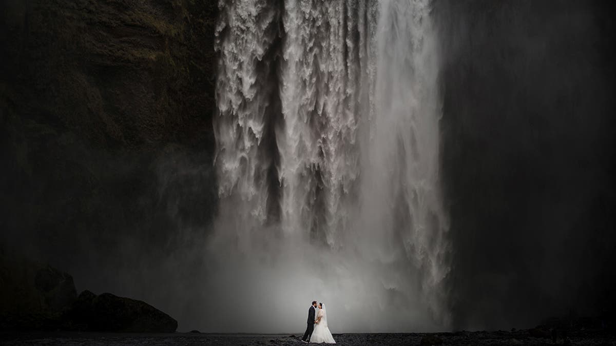 A wedding couple standing at the base of a massive, powerful waterfall, highlighting the epic scale of the landscape, shot by Susan Stripling.
